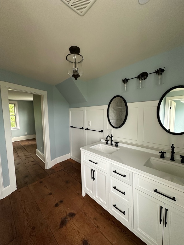 Light blue-painted bathroom with white dual-sink vanity, dark wood flooring, and cream-painted trim around doorways