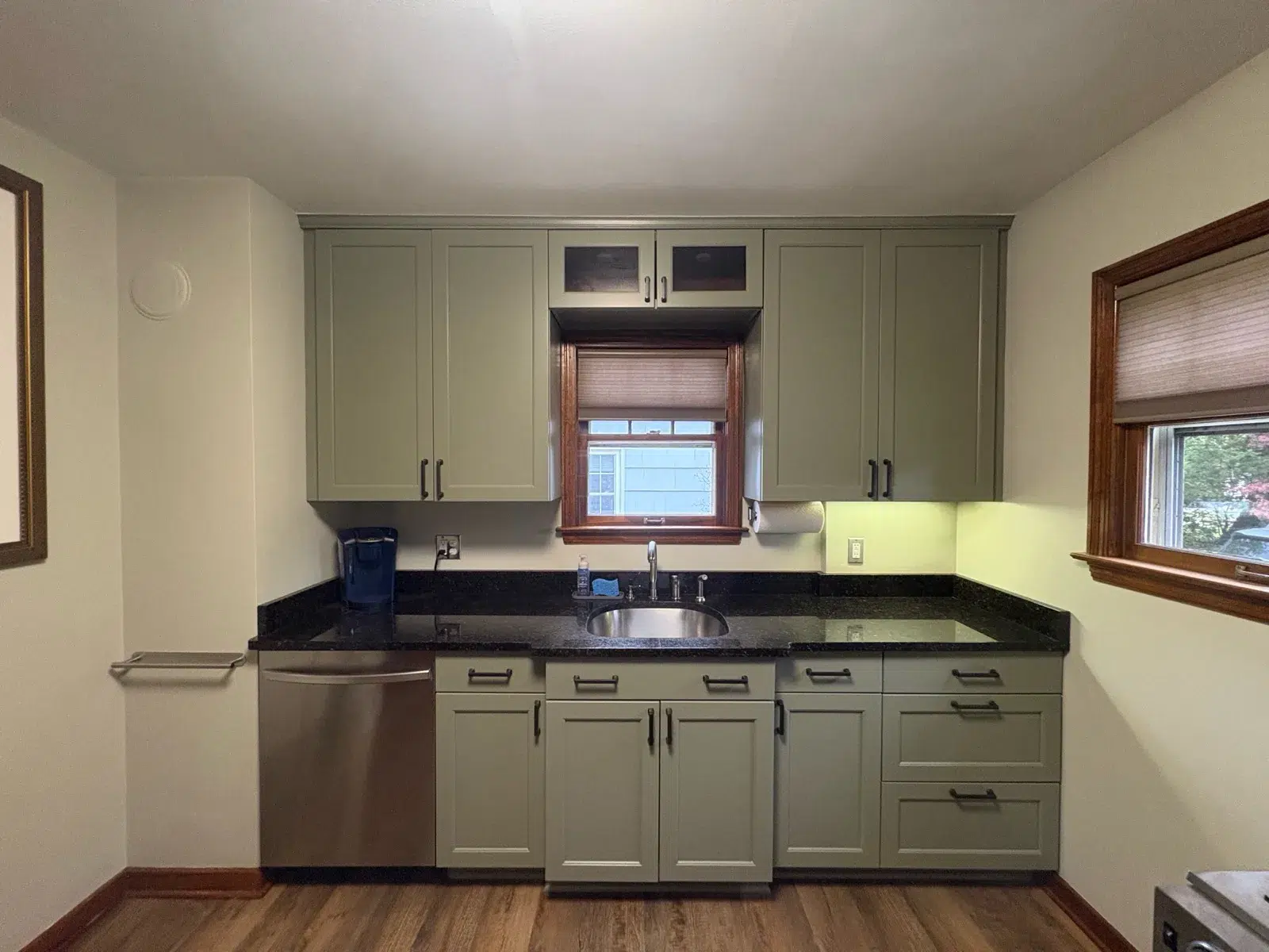 Sage Green Kitchen Cabinets Sage green-painted kitchen cabinets with black countertop, stainless steel sink and appliances, cream walls, and wood trim